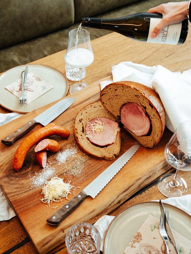 Angeschnittener Schinken im Brotteig, Käsekrainer und Wein auf einem Holzbrett